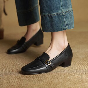Puede incluir: Mocasines negros de cuero con tac&oacute;n, puntera cuadrada y una correa en la parte superior asegurada con una hebilla dorada. Los zapatos tienen un tac&oacute;n bajo y cuadrado. La imagen muestra los zapatos con vaqueros azules.