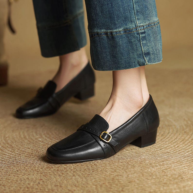 Puede incluir: Mocasines negros de cuero con tac&oacute;n, puntera cuadrada y una correa en la parte superior asegurada con una hebilla dorada. Los zapatos tienen un tac&oacute;n bajo y cuadrado. La imagen muestra los zapatos con vaqueros azules.