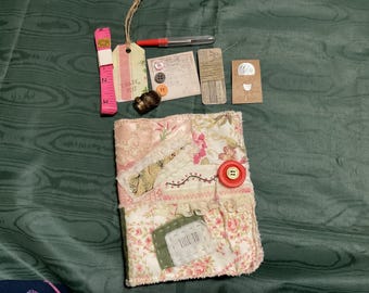 Puede incluir: Un diario de tela rosa y blanco con un botón rojo y el texto "Time To" en una etiqueta. El diario está rodeado de varios artículos de artesanía, incluyendo una cinta métrica rosa, una etiqueta marrón con el texto "Thank You", una pequeña bobina de hilo y algunos botones.