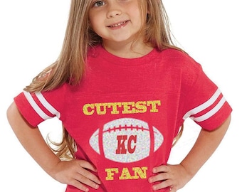 May include: A red and white striped football jersey with the words "Cutest KC Fan" in gold glitter lettering. The jersey features a white football with a gold "KC" inside.
