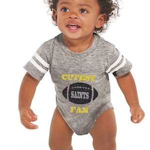 May include: A gray and white striped baby onesie with a black and yellow football graphic and the text "Cutest Saints Fan".