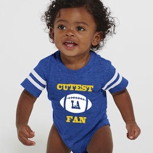 Puede incluir: Body de bebé azul con rayas blancas en las mangas. El body presenta el texto "Cutest LA Fan" y un gráfico de balón de fútbol blanco con "LA" en su interior. La tela tiene una textura jaspeada.