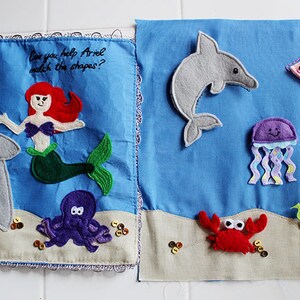 Op de afbeelding: Een viltboek voor kinderen met een zeemeermin, een schildpad, een dolfijn, een kwal, een krab, een vis en een schatkist. Het boek heeft de tekst "Can you help Ariel match the shapes?" op de omslag.