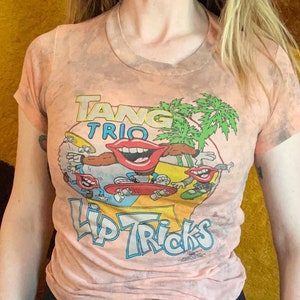 May include: Peach-colored t-shirt with a graphic featuring the text "Tang Trio Lip Tricks" and cartoon lips on skateboards. The shirt has a tie-dye effect and short sleeves. The model is wearing a black skirt.