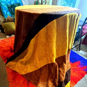 Op de afbeelding: Een ronde tafel bedekt met een veelkleurige, gestreepte deken. De deken heeft banden van geel, bruin en zwart. Een oranje shaggy vloerkleed is zichtbaar op de voorgrond.