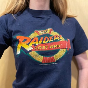 Può includere: T-shirt blu navy con il logo Atari Raiders of the Lost Ark in arancione, giallo e rosso. La maglietta vintage ha girocollo e maniche corte. La maglietta è in stile retrò.