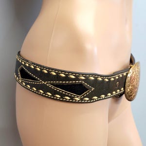 Pode incluir: Um cinto preto com uma fivela dourada decorativa. O cinto apresenta costuras creme e recortes em forma de diamante. A fivela tem um design ornamentado.