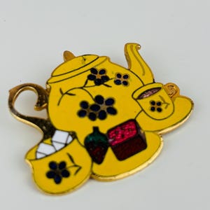 Puede incluir: Un broche de tetera de esmalte amarillo con un asa y borde dorados. El broche presenta una taza, cubos de azúcar y un pastel pequeño. Acentos florales azul oscuro están dispersos por la tetera y la taza.