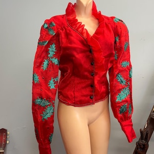 Könnte beinhalten: Eine rote, langärmelige Bluse mit Rüschenkragen und grüner Stechpalmen-Stickerei.