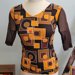 May include: A V-neck top with a geometric pattern in shades of brown, orange, and white. The top has sheer brown sleeves and a fitted silhouette. The design features squares, circles, and rectangles.