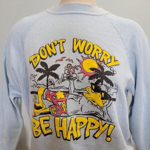 Puede incluir: Sudadera azul claro con la frase "DON'T WORRY BE HAPPY!" en amarillo. El gráfico presenta un pato de dibujos animados con gafas de sol, relajándose en una playa con palmeras y un sol. La sudadera tiene mangas largas.