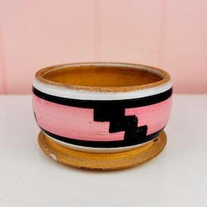 May include: A round ceramic planter with a matching saucer. The exterior features a wide horizontal pink band bordered by thin black lines, with a black stepped geometric pattern. The interior and base are a natural tan, unglazed clay color.