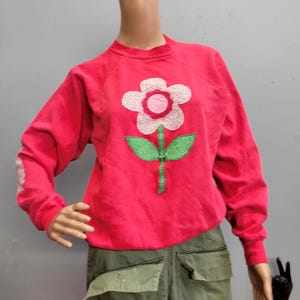 May include: A bright red sweatshirt with a large, appliqued flower design. The flower has white petals, a pink center, and green leaves and stem. The sleeves have a white design. The sweatshirt is displayed on a mannequin.