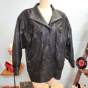 Peut inclure: Veste en cuir noir avec un motif texturé en forme de zèbre sur les épaules et le haut des manches. La veste a une fermeture boutonnée, deux poches et un col classique. La veste est présentée sur un mannequin.