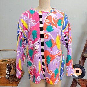 Könnte beinhalten: Ein langärmliges T-Shirt mit einem lebendigen, abstrakten Muster in Lila, Rosa, Gelb und Blau. Das Design umfasst skurrile Formen und einen schwarz-weißen vertikalen Streifen. Das Shirt wird auf einer Schaufensterpuppe präsentiert.