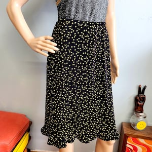 May include: A black midi skirt with a repeating pattern of yellow crescent moons and stars. The skirt has a ruffled hem and is paired with a silver top. The skirt is knee-length and appears to be made of a lightweight fabric.