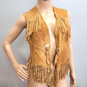 May include: A tan suede vest with fringe detailing. The sleeveless vest features fringe along the bottom and shoulders. It has two silver-tone metal clasps and a stitched design. The vest is displayed on a mannequin.