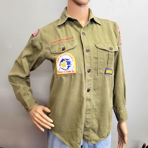 May include: Olive green long-sleeve shirt with button-down front and two chest pockets. The shirt features patches with text and graphics, including "Twin Valley Council Scout-O-Rama '82" and an American flag patch. The shirt is displayed on a mannequin.