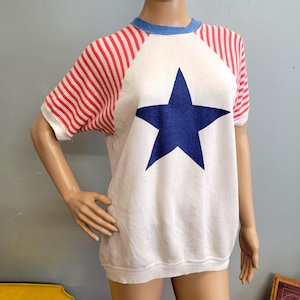 May include: White short-sleeved shirt with a large blue star in the center. The sleeves are red and white striped. The shirt has a blue crew neck. The shirt is displayed on a mannequin.