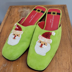 May include: A pair of lime green slip-on shoes with a Santa Claus design. Each shoe features a sequined Santa face and a red and black insole with the brand name. The shoes are on a wooden surface.