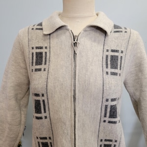 Könnte beinhalten: Hellgraue Strickjacke mit Reißverschluss, Kragen und langen Ärmeln. Die Strickjacke hat ein geometrisches Muster in Dunkelgrau. Der Reißverschluss hat einen dekorativen silbernen Zipper. Die Strickjacke wird auf einer Schaufensterpuppe präsentiert.