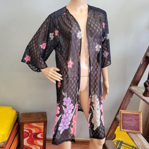 May include: Black, open-front kimono-style robe with a floral pattern in pink, purple, and white. The sheer fabric has a subtle geometric texture. The robe has three-quarter sleeves and falls to the ankles.