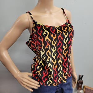 May include: Black camisole top with a flame pattern in red, orange, and yellow. The tank top has thin spaghetti straps and a scoop neckline. The flame design covers the entire top, creating a bold and eye-catching look. The top is paired with blue jeans.