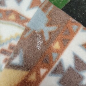 K&ouml;nnte beinhalten: Nahaufnahme eines gefalteten Fleece-Stoffs mit geometrischem Muster. Das Design zeigt braune, wei&szlig;e, hellblaue und gelbe Formen. Der Stoff hat ein weiches, strukturiertes Aussehen, das W&auml;rme und Komfort suggeriert. Das Muster erinnert an s&uuml;dwestliche oder indianische Designs.