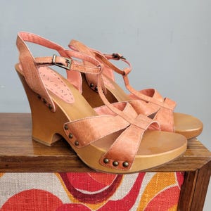 Puede incluir: Un par de sandalias de cuña de cuero color melocotón con diseño en T, correas de tobillo ajustables con hebillas de metal y suelas de madera. Los laterales de las correas de cuero están fijados a la base de madera con tachuelas metálicas.