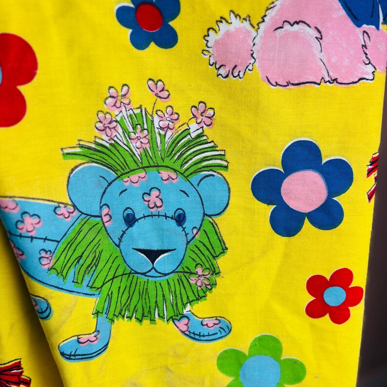 May include: Bright yellow fabric featuring a repeating pattern of cartoon lions, flowers, and a dog. The lion is blue with a green mane and pink flowers. The flowers are blue, red, and green.