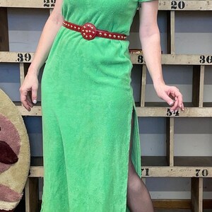 Puede incluir: Un vestido maxi verde con capucha, una abertura lateral y un cintur&oacute;n rojo con tachuelas. El vestido est&aacute; hecho de un tejido suave de toalla.