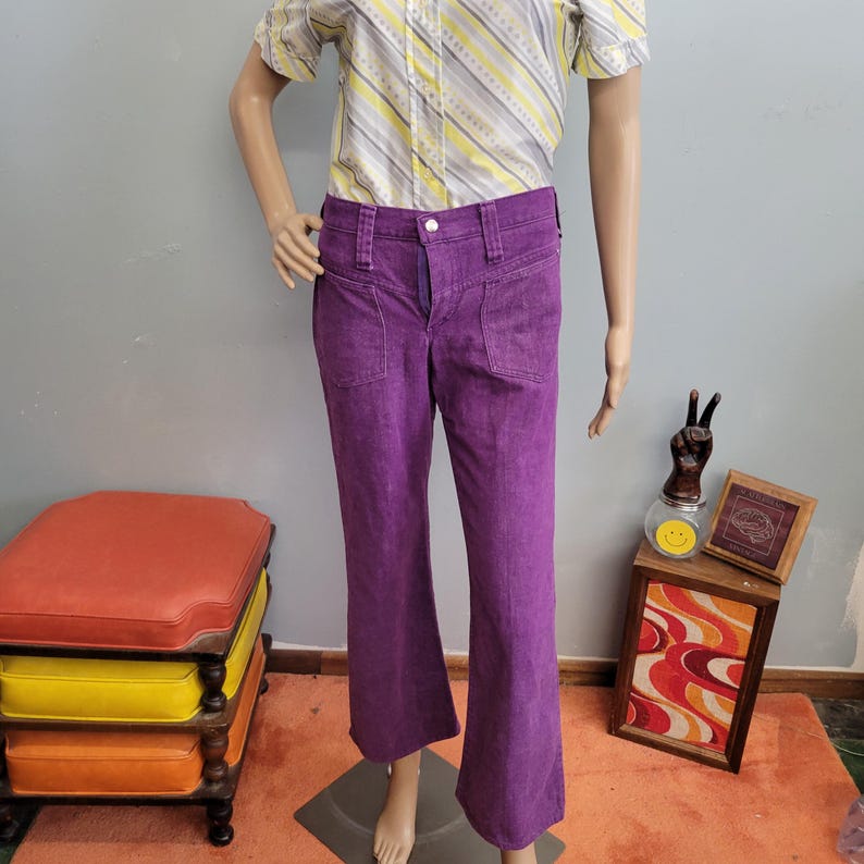 May include: A pair of vibrant purple flared pants with front pockets. The pants are displayed on a mannequin, showcasing their retro style. The fabric appears to be a soft, textured material. The pants are paired with a striped button-up shirt.