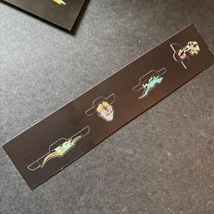 以下が含まれることがあります： 黒いカードに4つのエナメルピン。ピンは、デザインのある帽子、トカゲ、頭蓋骨、生き物を描いています。ピンには、金、青、ピンク、黄色のアクセントがあります。