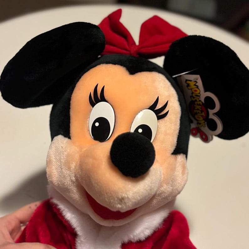 Pode incluir: Um boneco de pel&uacute;cia da Minnie Mouse com orelhas pretas, um la&ccedil;o vermelho e uma roupa vermelha e branca. A boneca tem olhos brancos grandes, c&iacute;lios pretos e um nariz preto. Uma etiqueta est&aacute; presa &agrave; orelha da boneca.