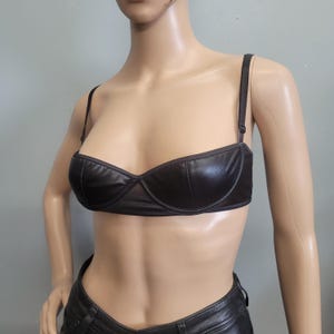 Può includere: Un bralette in similpelle marrone scuro caratterizzato da un design strutturato con ferretto, spalline sottili regolabili e una finitura liscia. Il capo presenta uno scollo dritto e una silhouette corta che cade sopra la vita.