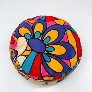 Pode incluir: Uma bolsa redonda com fecho de correr apresentando um padrão floral multicolorido vibrante com formas arrojadas em tons de roxo, amarelo, vermelho, rosa e turquesa. O tecido tem uma textura suave e um fecho dourado percorre a borda lateral.