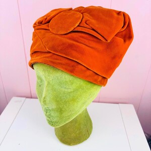 May include: A burnt orange velvet turban-style hat featuring a gathered, pleated design and a prominent circular fabric accent on the front. The hat is displayed on a textured, light green mannequin head, showcasing the soft, plush fabric texture.