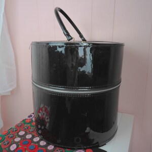 May include: A black, cylindrical, patent leather handbag with a silver zipper and a black handle. The bag is sitting on a patterned surface. The bag is a vintage style.