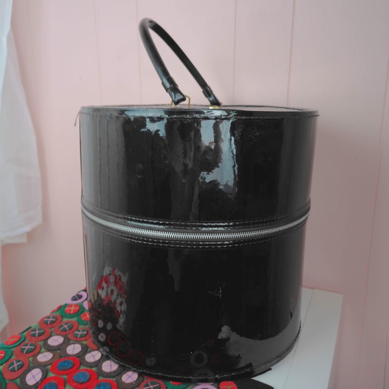 May include: A black, cylindrical, patent leather handbag with a silver zipper and a black handle. The bag is sitting on a patterned surface. The bag is a vintage style.