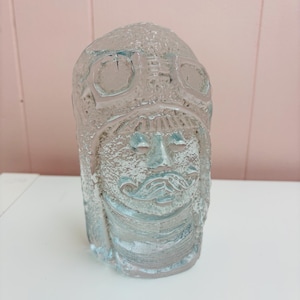 Puede incluir: Una escultura de vidrio azul claro texturizado de un hombre barbudo con casco. La escultura tiene forma rectangular con rasgos faciales detallados y diseño de casco. El vidrio tiene un aspecto esmerilado.