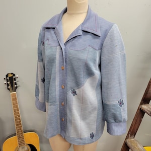 May include: A vintage blue button-down shirt with a patchwork design. The shirt features a collared neckline, long sleeves, and small floral embroidery. The shirt has a relaxed fit and is made of a soft, textured fabric.