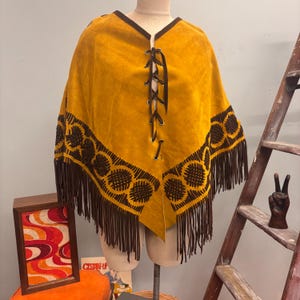 May include: Mustard yellow poncho with brown fringe and decorative stitching. The poncho has a V-neck with a brown lace-up closure. The bottom edge features a repeating circular pattern and long fringe.