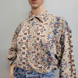 May include: Beige button-down shirt with a Southwestern-inspired pattern in blue, red, and black. The long-sleeved shirt features a chest pocket and a classic collar. The shirt is displayed on a mannequin.