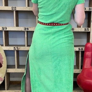 Puede incluir: Un vestido largo de toalla verde con capucha y cintur&oacute;n rojo con tachuelas. El vestido tiene una abertura lateral y se lleva con tacones negros.