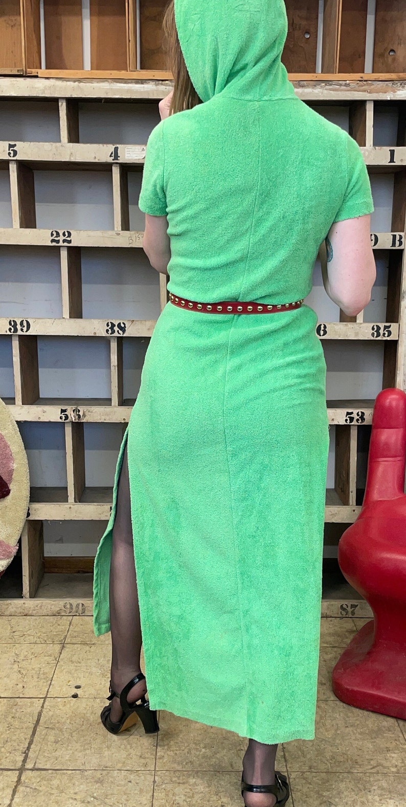Puede incluir: Un vestido largo de toalla verde con capucha y cintur&oacute;n rojo con tachuelas. El vestido tiene una abertura lateral y se lleva con tacones negros.
