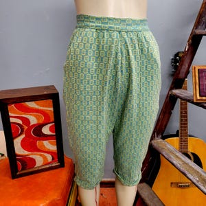 May include: Vintage teal and yellow patterned capri pants. The capris feature a high waist, pleats, and a rolled hem. The fabric has a repeating geometric design. The pants are displayed on a mannequin.
