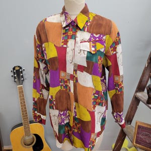 Peut inclure: Une chemise à manches longues avec un motif patchwork dans les tons de marron, orange, jaune, violet et rouge. La chemise présente un col, une poche poitrine et des poignets boutonnés. Le tissu a un aspect soyeux.