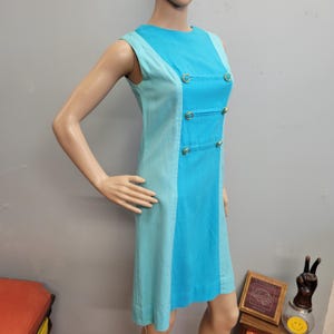 May include: A sleeveless, knee-length dress in shades of turquoise and light blue. The dress features a color-blocked design with a central panel of bright turquoise and side panels of light blue. Gold-colored circular accents adorn the front.