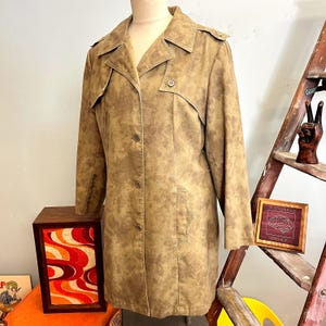 May include: A tan vintage coat with a button-up front, lapel collar, and shoulder epaulets. The coat has a mottled pattern and a tailored fit. The coat is displayed on a mannequin. A wooden box with an abstract design is in the background.