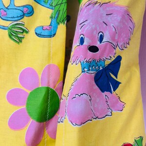 May include: Bright yellow fabric featuring a repeating pattern of pink cartoon dogs with blue bows, blue eyes, and black noses. Large pink flowers with green centres and cartoon figures with blue feet and green arms are also visible.